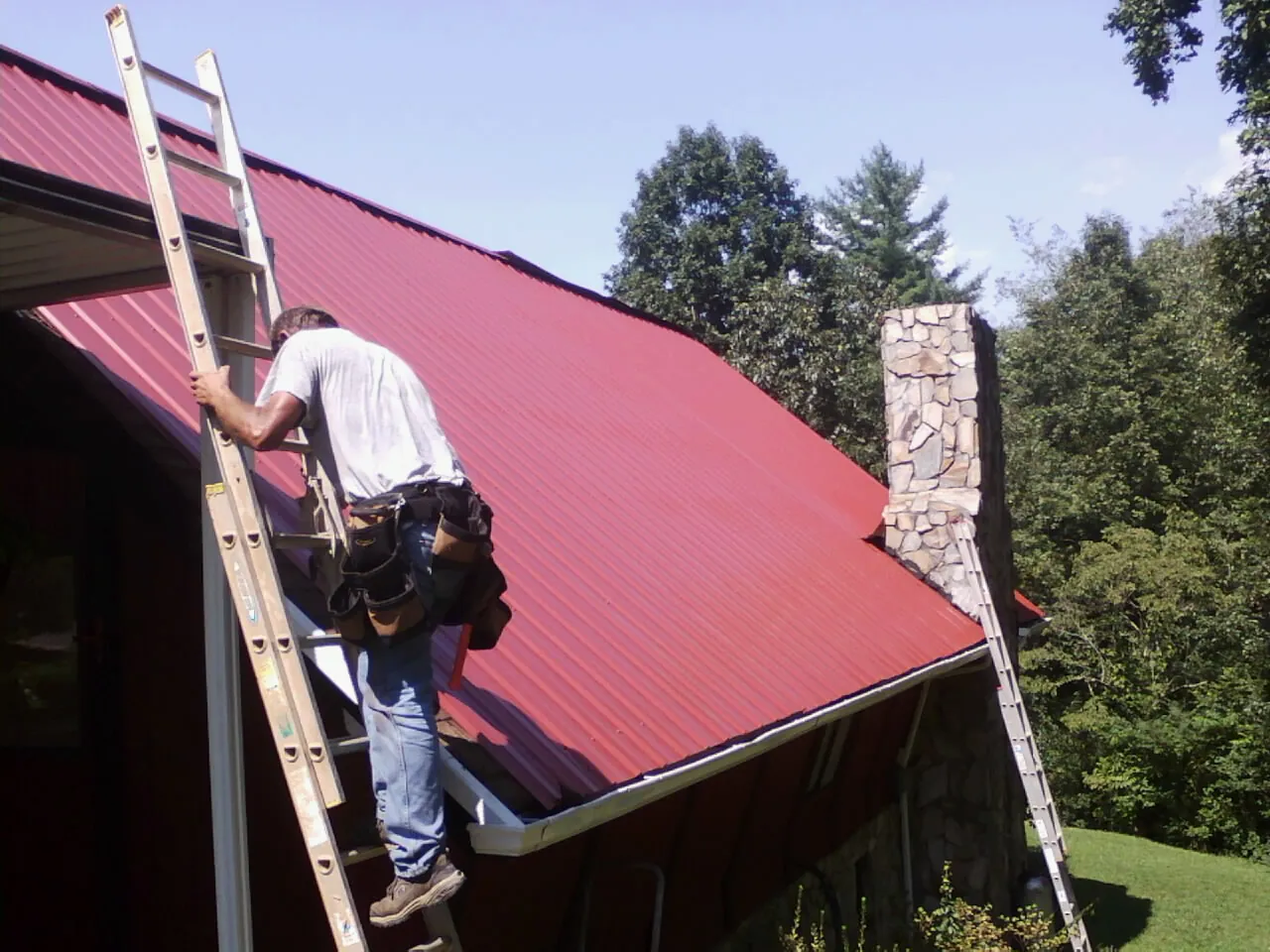 Roofer on a ladder beside a red metal roof and stone chimney — Soffit & Fascia Repair by Roofing Brothers in Flossmoor